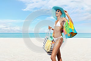 Happy woman at the beach side wearing a bikini, sunglasses and blue sun hat holding beach bag and towel in a sunny day with blue