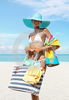 Happy woman at the beach side wearing a bikini, sunglasses and blue sun hat holding beach bag and towel in a sunny day with blue