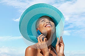 Happy woman at the beach side wearing blue sun hat and using mobile phone in a sunny day with blue sky. Concept of summer beach