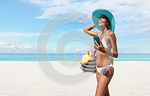 Happy woman at the beach side wearing sun hat, holding a beach bag and using mobile phone in a sunny day with blue sky. Concept of