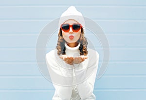 Happy woman blowing red lips sends air kiss wearing a heart shape sunglasses, knitted hat, sweater over blue