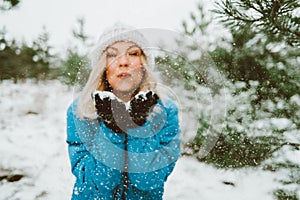 Happy woman blowing snow from palms with mittens to camera on winter forest background. Portrait of cute girl childishly photo