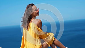 Happy woman in bright yellow dress and red sunglasses sitting on cliff edge against amazing seascape.