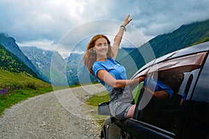 Happy woman in a car driving on the road against a background of mountains, light hair color, raises her hand up. Summer