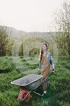A happy woman with a cart works in her country home in the countryside against a backdrop of green grass and sunset sun