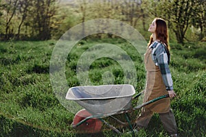 A happy woman with a cart works in her country home in the countryside against a backdrop of green grass and sunset sun