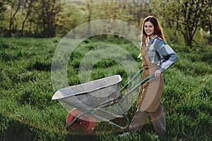 A happy woman with a cart works in her country home in the countryside against a backdrop of green grass and sunset sun