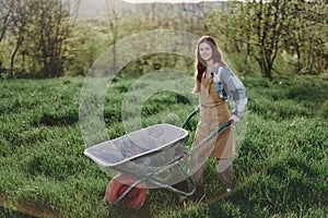 A happy woman with a cart works in her country home in the countryside against a backdrop of green grass and sunset sun