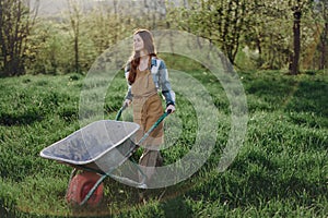 A happy woman with a cart works in her country home in the countryside against a backdrop of green grass and sunset sun