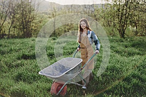 A happy woman with a cart works in her country home in the countryside against a backdrop of green grass and sunset sun