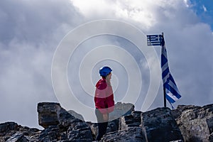 Happy woman with climbing equipment on cloud covered mountain summit of Mytikas Mount Olympus, Greece. Greek flag on top