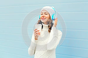 Happy woman with coffee cup enjoys listens to music in headphones wearing a knitted hat, sweater over blue