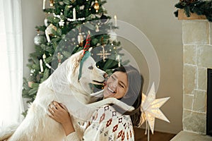 Happy woman in cozy sweater hugging cute white dog in festive accessory on background of stylish christmas tree. Winter holidays.