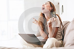 Happy woman with credit card and laptop sitting on sofa