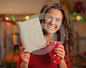 Happy woman with cup of hot chocolate reading book in kitchen