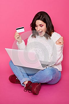 Happy woman with dark hair clenching fists and yelling happily, holding credit in hands, having success online purchase, lady