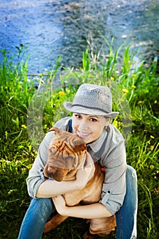 Happy woman in denim overalls and hat with her dog Shar Pei sitting in the meadow near the lake at sunset