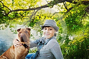 Happy woman in denim overalls and hat with her dog Shar Pei sitting in the meadow near the lake at sunset