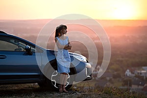 Happy woman driver in blue summer dress enjoying warm evening near her car. Travel and vacations concept