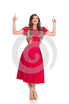 Happy Woman In Elegant Red Dress And Gold High Heels Is Looking Up, Pointing And Smiling