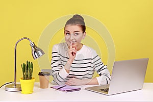 Happy woman employee at workplace shushing with silence gesture, asking to keep secret.