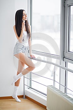 Happy woman enjoying homey morning. Attractive young woman standing near the window, drinking a tea or coffee from white