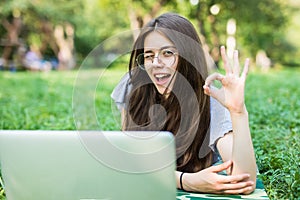 Happy woman in eyeglasses lying on grass in park with laptop computer and showing ok sign