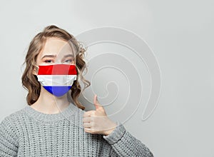 Happy woman in face mask holding thumb up with Netherlands  flag background. Flu epidemic and virus protection concept