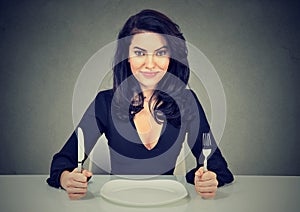 Happy woman with fork and knife sitting at table with empty plate