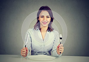Happy woman with fork and knife sitting at table with empty plate