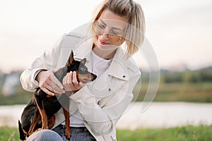 Happy woman with glasses walking on grass in park, hugging her small companion dog. Dog owner pets his pet during a