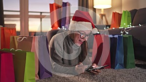 Happy woman with glasses wearing a santa claus hat is lying on the carpet and makes an online purchase using a credit