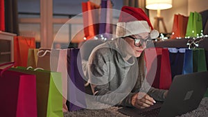 Happy woman with glasses wearing a Santa Claus hat is lying on the carpet and makes an online purchase using a credit