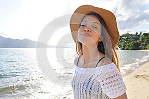 Happy woman with hat breathing and smiling at the beach on summer vacation. Beautiful young female with closed eyes enjoy nature