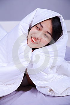 Happy woman hiding under blanket on bed in bedroom
