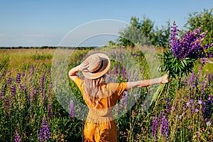 Happy woman holding bouquet of lupin flowers walking in summer meadow. Girl opens arms enjoying landscape