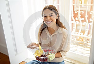 Happy woman holding bowl of fruits, enjoying a healthy snack by window