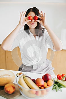 Happy woman holding cherry tomato at eyes and smiling in modern kitchen. Having fun making salad