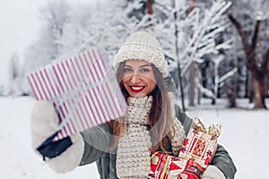 Happy woman holding Christmas presents gift boxes in snowy winter park offering one. Festive holiday season. Space