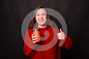 A happy woman is holding a cup of hot drink and laughing at camera is showing a thumb up