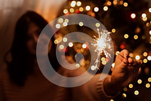 Happy woman holding firework at christmas tree with golden lights. Stylish girl with burning sparkler celebrating in festive dark
