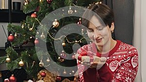 Happy woman is holding little Christmas gift in hands sitting near New Year tree. Smiling beautiful female in Christmas red sweate