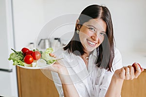 Happy woman holding plate with fresh lettuce, cherry tomatoes, arugula in kitchen. Healthy eating