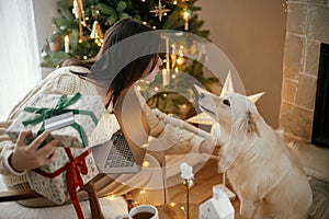 Happy woman holding stylish christmas gifts with credit card and caressing cute white dog in festive decorated christmas room with