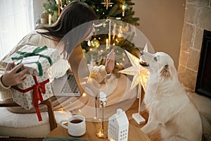 Happy woman holding stylish christmas gifts with credit card and playing with cute white dog in festive decorated christmas room