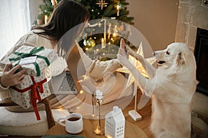 Happy woman holding stylish christmas gifts with credit card and playing with cute white dog in festive decorated christmas room