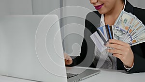 Happy woman holding US dollars and credit cards near her laptop