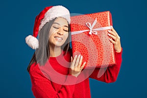 Happy woman holds gently long awaited christmas gift. Beautiful woman in santa claus hat on blue background Xmas concept