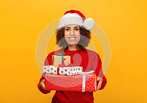 Happy woman holds in hands stack of christmas gifts. female in santa claus hat on orange background, smiling. Xmas