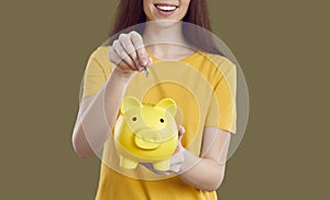 Happy woman holds yellow piggy bank, throwing coin into it stands on solid green background